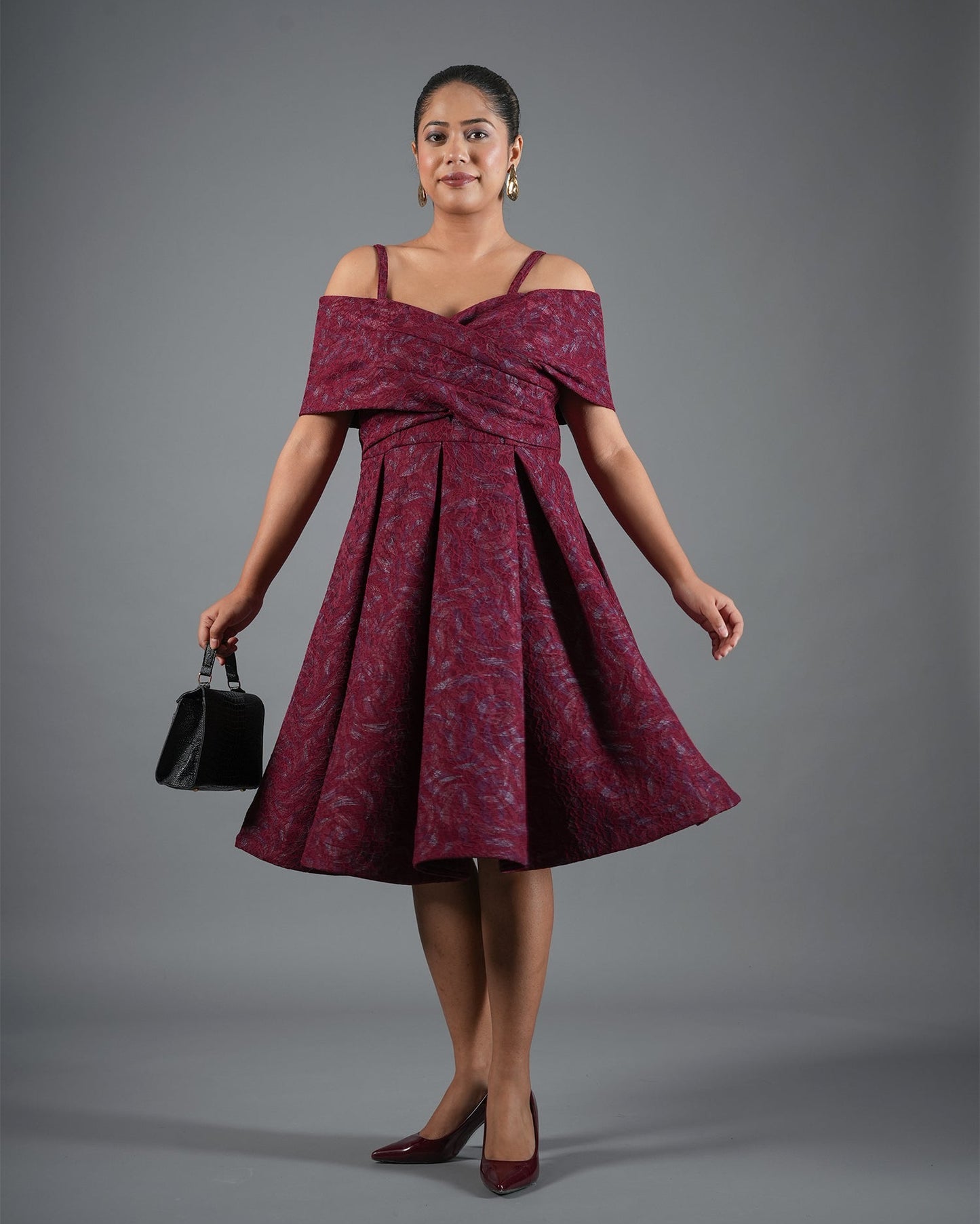Woman wearing a burgundy off-shoulder dress holding a black handbag against a gray background