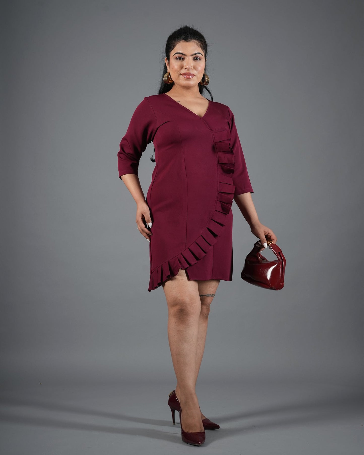 Woman wearing a burgundy dress holding a matching handbag against a gray background
