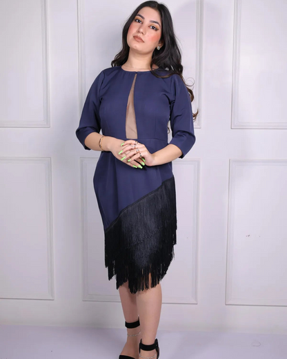 Woman wearing a navy blue top with a black fringed skirt against a white paneled wall.