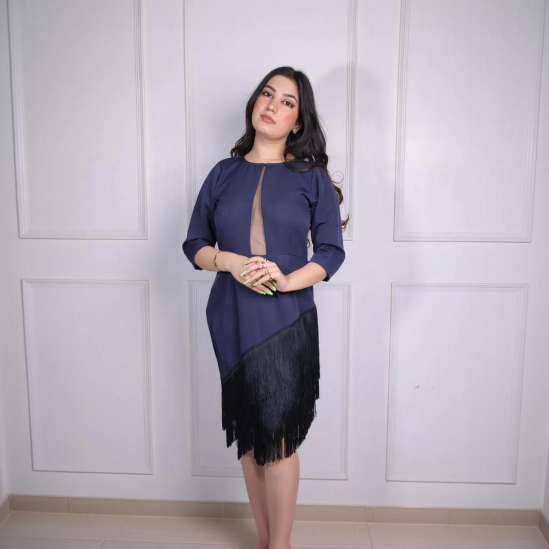 Woman wearing a navy blue top with a black fringed skirt against a white paneled wall.