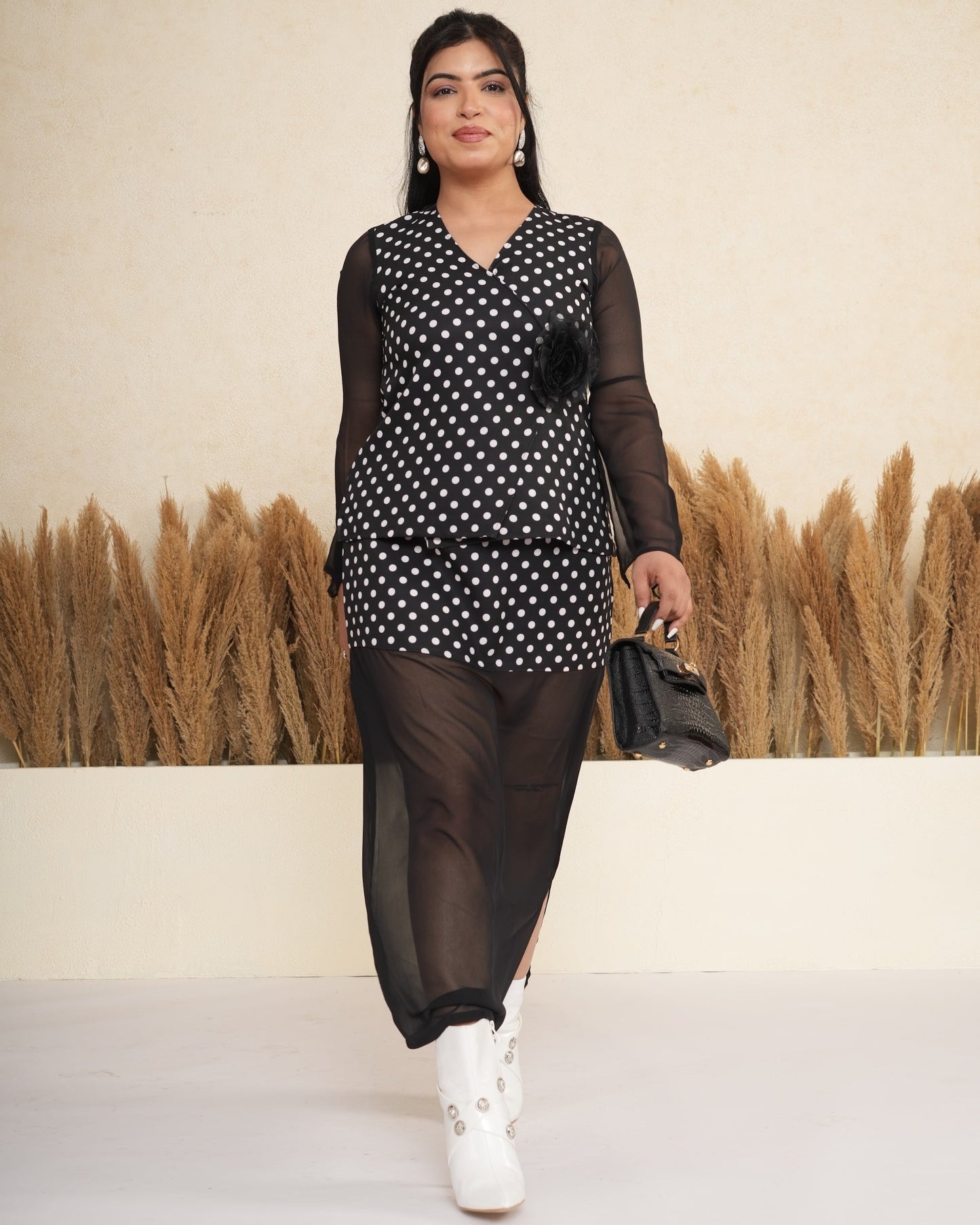 Woman in a black and white polka dot dress with sheer sleeves and black pants, standing against a beige wall with potted plants.