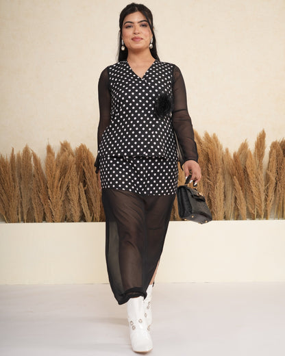 Woman in a black and white polka dot dress with sheer sleeves and black pants, standing against a beige wall with potted plants.