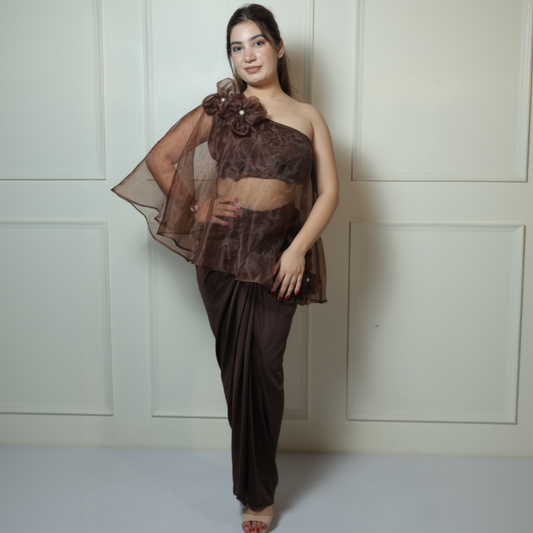 Woman wearing a brown floral one-shoulder top and dhoti pants against a white paneled wall.