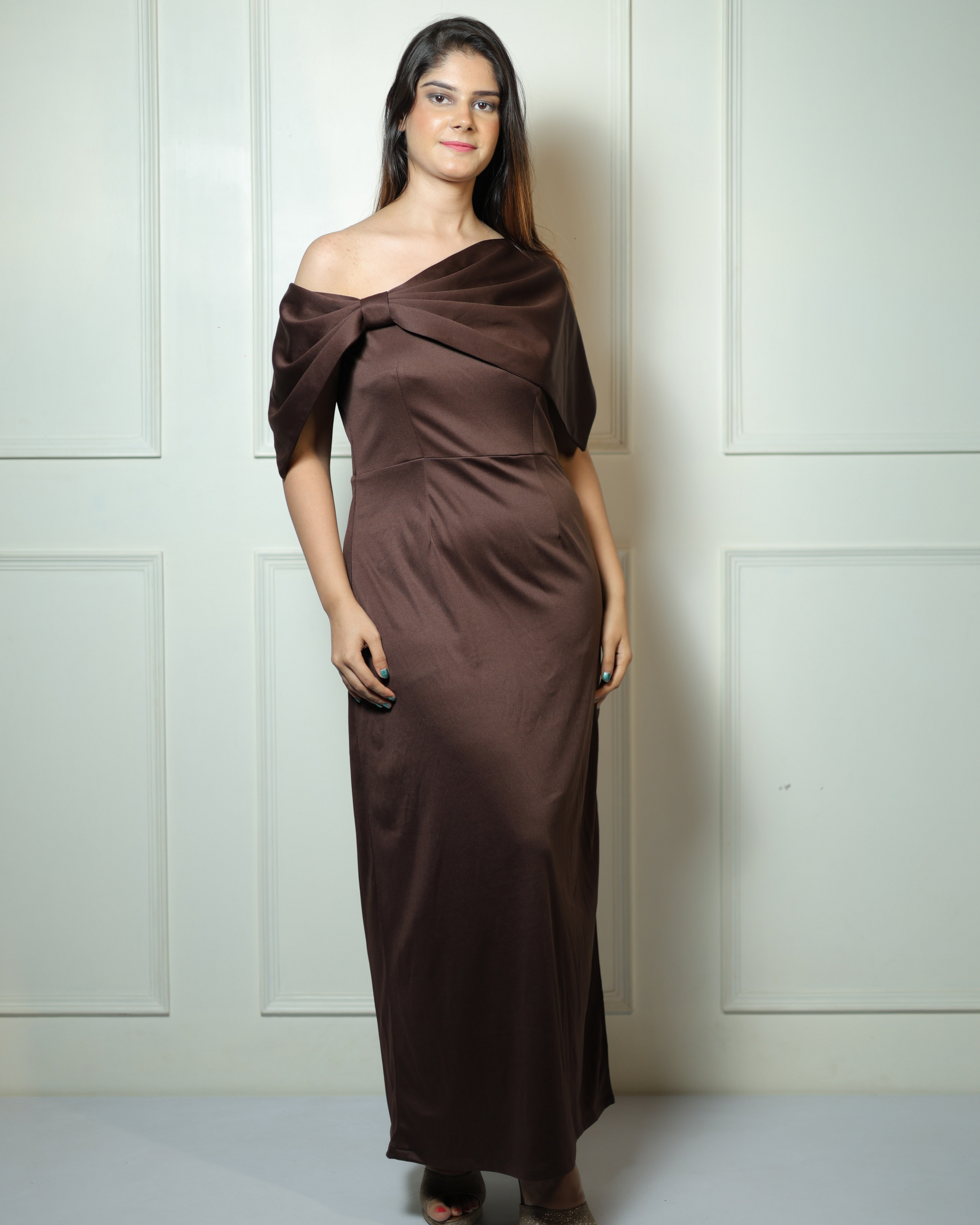 Woman wearing a brown off-shoulder dress against a white paneled wall.