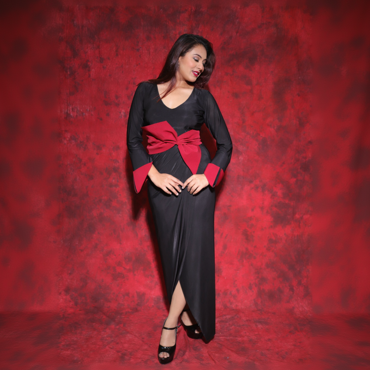 A woman posing in a black lycra draped dress with red cuffs and a detachable bow belt at the back, standing against a red backdrop.